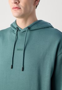 Felpa con cappuccio in misto cotone color teal, dotata di tasca a marsupio e cordini con punte nere. Logo "BOSS" ricamato sul petto.