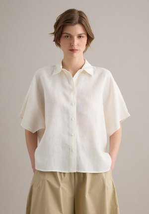 Giovane donna con capelli corti che indossa una camicia bianca ampia a maniche corte con bottoni e pantaloni beige, in piedi davanti a uno sfondo uniforme.