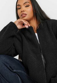 Czarna polarowa bomberka z zamkiem błyskawicznym i teksturowanym materiałem. Posiada okrągły dekolt i luźny krój, zestawiona z ciemnymi dżinsami.