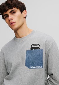 Felpa grigia con scollo rotondo, caratterizzata da una tasca in denim blu e una grafica di occhiali da sole neri. Testo "KARL LAGERFELD" sulla tasca.