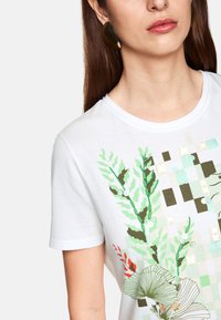 White cotton t-shirt featuring a green plant print with red accents. Includes decorative beads and a geometric background pattern.