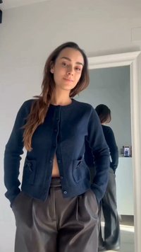 Frau mit langen Haaren, die eine marineblaue Strickjacke und lockere schwarze Hosen trägt, steht mit den Händen in den Taschen vor einem Spiegel, der ihren Rücken reflektiert.