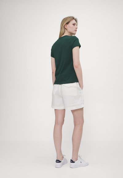 Mørkegrønn bomull t-skjorte, kort erm, kombinert med hvite linshorts som har en lomme på baksiden. Modellen har på seg hvite sko med mørke detaljer.