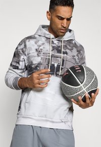 Grå og hvid hættetrøje med abstrakt print, snor og lommer foran, båret af en mand der holder en sort og grå tekstureret basketball.