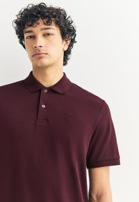 Vínová polokošile z hladké tkaniny s límečkem, tříknoflíkovým zapínáním a vyšitým logem na hrudi. Krátké rukávy.