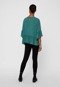 Top teal morbido con maniche arricciate e dettaglio a chiave nella parte posteriore, abbinato a jeans skinny neri e stivaletti.