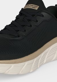 Μαύρο αθλητικό παπούτσι Skechers BOBS με υφασμάτινο επάνω μέρος, μαύρα κορδόνια, μπεζ ενδιάμεση σόλα και λευκή εξωτερική σόλα με σχέδιο, εμφανίζεται σε κοντινό πλάνο.