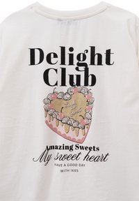 Camiseta de algodón blanca con un gráfico de un pastel en forma de corazón con acentos rosas, texto brillante y tipografía negra. El texto dice "Delight Club."