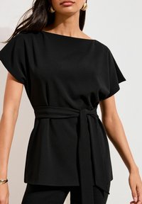 Sort, kortærmet bluse med en bred halsudskæring, taljen er indsnøret og bundet med et bælte, og stoffet er glat og tekstureret.