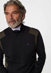 Mogen man med grått hår och skägg, iklädd en svart och olivgrön ziptröja över en vit höghalspolo, som tittar åt vänster.