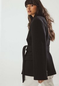 Must bleiser kohandatud lõikega, laia krae ning vöökinnitusega. Omaduseks on tekstureeritud kangas ja nöörideemaldused varrukaotstel. Koos valgete pükstega.