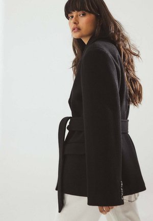 Zwarte blazer met een getailleerde vorm, brede revers en een ceintuur met strik. Voorzien van een gestructureerde stof en knoopdetails op de mouwen. Gecombineerd met witte broek.