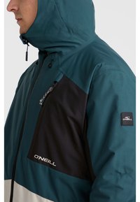 Chaqueta color bloqueada en azul verdoso y negro, con un bolsillo de pecho con cremallera, capucha ajustable y parche del logo de O'Neill en la manga.
