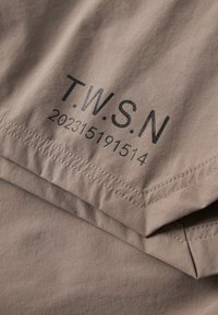 Nahaufnahme eines beigen Stoffs mit schwarzem Text "T.W.S.N" und den Zahlen "202315191514" über genähten Nahtlinien gedruckt.
