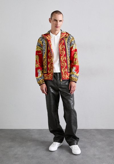 Man som st�år klädd i en färgglad mönstrad jacka med djurtryck, vit polo-tröja, svarta strukturerade byxor och vita sneakers framför en neutral vägg.