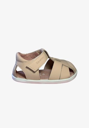 Sandalia beige de cuero para niño pequeño con tira ajustable de velcro y diseño de punta abierta sobre suela blanca de goma.