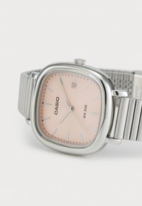 Montre Casio avec un boîtier carré en argent, un cadran rose, des aiguilles argentées et un affichage de la date. Le bracelet en métal présente des rainures horizontales et est étanche jusqu'à 50m.