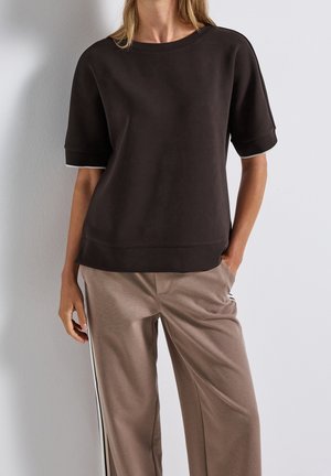 Femme portant un haut ample à manches courtes marron foncé et un pantalon beige à jambes larges avec des bandes latérales noires et blanches, debout avec une main dans la poche.
