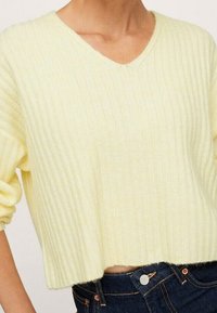 Maglione a coste di un giallo chiaro con scollatura a V e design cropped, caratterizzato da una texture morbida e spalle basse, abbinato a jeans in denim scuro.