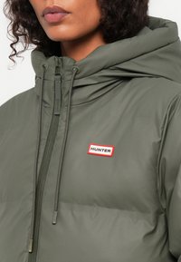 Maslinasto zelena jakna s puhovima s visokim ovratnikom, patentnim zatvaračem i podesivom kapom. Sadrži crveno-bijelu oznaku "HUNTER". Glatka tekstura tkanine.