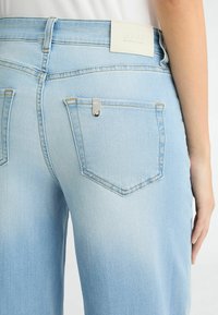 Un jean en denim bleu clair présente une taille mi-haute, des coutures visibles et un petit accent métallique sur la poche arrière. L'étiquette indique "LIU-JO."