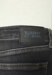 Mustat farkut, joissa on tekstuurilla varustettu nahkapatchi, jossa lukee "TOMMY JEANS" kohokuvioituna. Ominaisuuksina näkyvät ompeluyksityiskohdat.