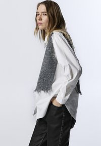 Gilet grigio lavorato a maglia con bordi sfrangiati indossato sopra una camicia bianca con colletto, abbinato a pantaloni neri con dettagli di cucitura a contrasto.