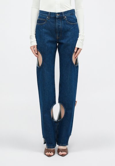 Vaqueros de mezclilla azul con grandes recortes en los muslos, silueta de pierna recta y detalles sutiles de costura. Combinados con tacones abiertos marrones.