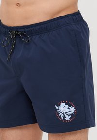Tamnoplave kupaće gaće s elastičnom strukom i crnim vezicama. Sadrže cvjetni print logotipa u bijeloj i narančastoj boji na lijevom bedru.