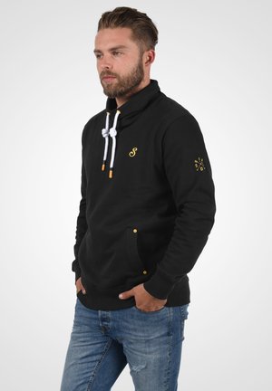Sudadera negra con cuello alto, cordones blancos, detalles amarillos y bolsillos frontales. Tela texturizada y sutil marca en el pecho.
