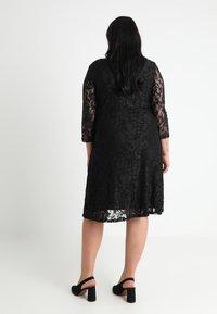 Svart spetsklänning med trekvartsärmar och utsvängd nederkant, prydd med blommönster. Bärs med svarta högklackade skor.