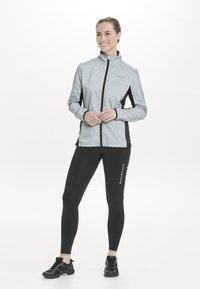 Lys grå jakke med sorte detaljer, høj krave og frontlynlås, parret med sorte leggings med et hvidt logo og sorte træningssko.