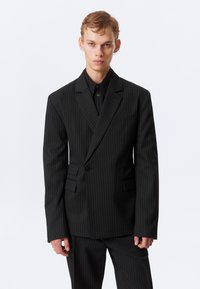 Svartruteret dressjakke med en enkelt knapp, klafferevers og to frontlommer. Båret over en svart skjorte, det ser glatt ut.