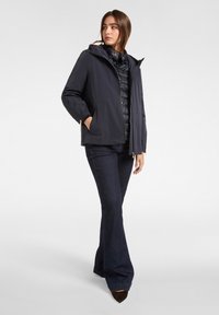 Giacca impermeabile blu navy con imbottitura interna trapuntata a contrasto, cappuccio regolabile e tasche con cerniera, abbinata a jeans scuri a zampa.