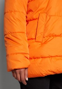Chaqueta acolchada naranja con diseño de rombos, que cuenta con un bolsillo lateral y un tejido grueso e aislante; la manga presenta una textura suave.