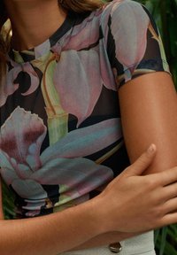 Košile s květinovým vzorem je v kombinaci růžových a zelených odstínů s jemnou texturou; má krátké rukávy a kulatý výstřih.