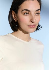 Artimas plano kadras, kuriame matomas žmogus, vilkintis šviesiai smėlio spalvos, prigludusią palaidinę su apvaliu iškirpimu. Akį traukia auksinės žiedinės auskarai ir lygi odos tekstūra.