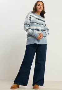 Maglione a maglia azzurro chiaro con motivi geometrici bianchi e neri, polsini a coste, abbinato a jeans in denim scuro a gamba larga e zoccoli in suede marrone.