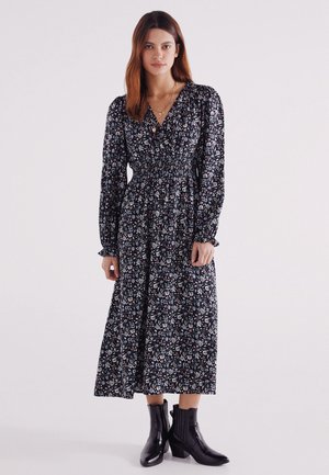 Schwarzes Blumenkleid mit langen Ärmeln und taillierter Taille, mit bunten Mustern, kombiniert mit schwarzen Stiefeletten.