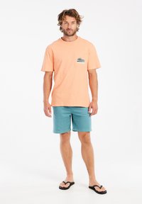 Korte mouwen perzikkleurig T-shirt met een golfgrafiek op de borstzak, gecombineerd met teal korte broek en zwarte flip-flops. De stof lijkt zacht en casual.