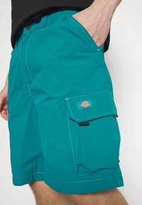 Shorts cargo bleu sarcelle en tissu léger, avec une poche latérale à rabat et des coutures blanches contrastantes.