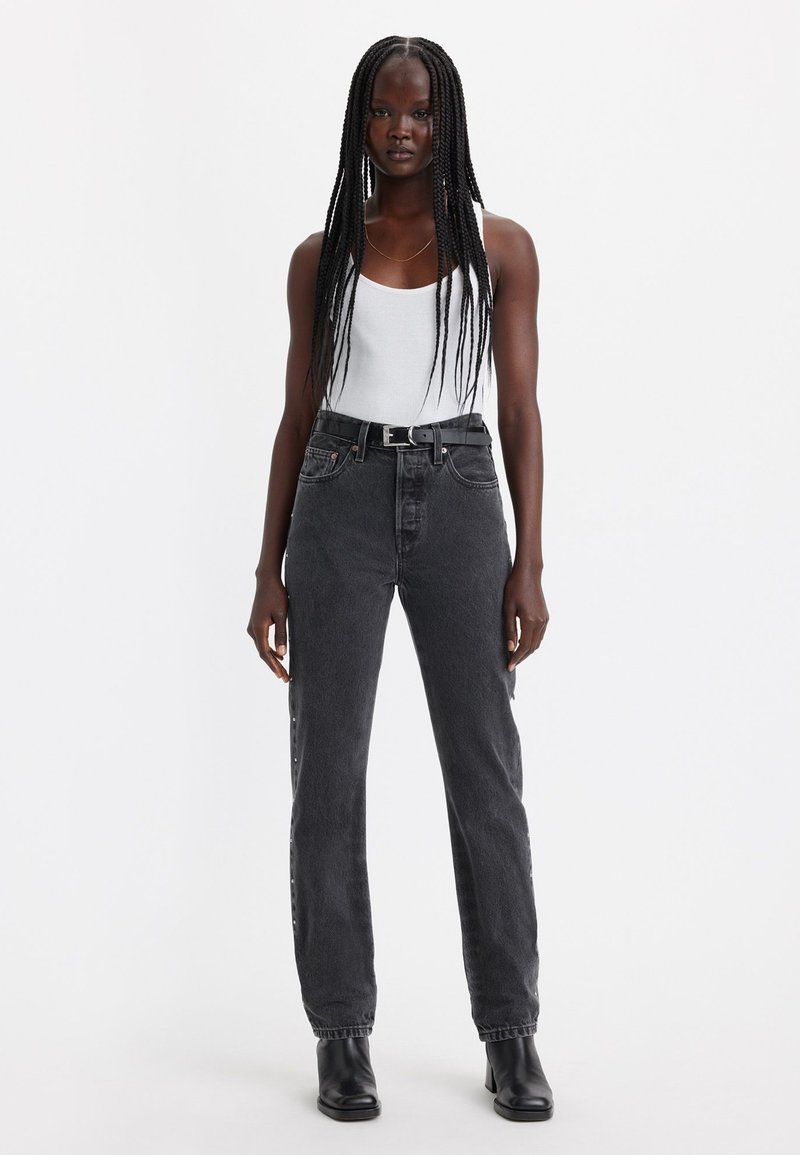 Jeans noirs avec une coupe droite, dotés de cinq poches et de boutons sur les côtés, associés à un débardeur blanc et des bottes à talons noires.