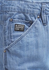 Ljusblå denimjeans med en framficka som har en G-Star Originals-etikett. Synlig sömnad och en metallknappdetalj.