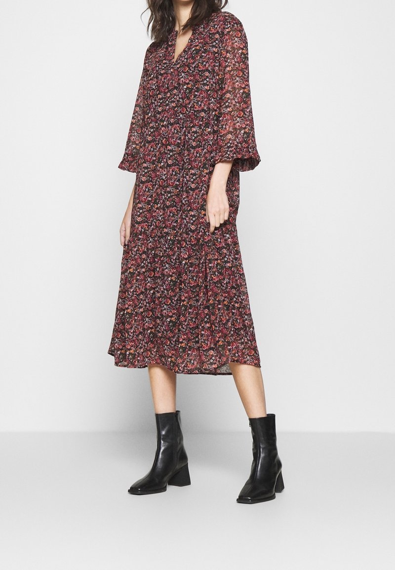 Femme portant une robe midi fleurie sombre à manches trois-quarts et des bottines noires à talons moyens, debout devant un fond uni.