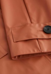 Gros plan d'une manche de veste en cuir brun avec une sangle cousue et un grand bouton noir en détail de fermeture.