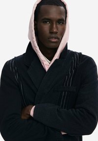 Abrigo negro con detalles de rayas y cuello de pico, superpuesto a una sudadera con capucha de color rosa claro. Tejido texturizado con un ajuste relajado.