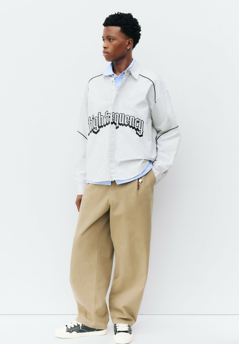 Jeune personne aux cheveux bouclés foncés, portant un pantalon large beige, des baskets blanches et une chemise à carreaux claire avec le texte "high frequency" sur la poitrine.