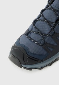 Sapatilha de caminhada masculina com parte superior em malha na cor azul-marinho, sola de borracha texturizada preta e atacadores refletivos. O design inclui sobreposições de suporte.