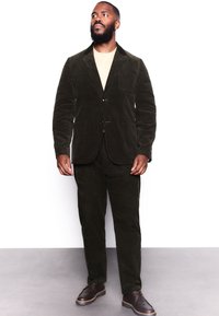 Costume en velours côtelé vert foncé avec une veste à simple boutonnage, des revers à cran, et un pantalon assorti. Des chaussures marron complètent le look.