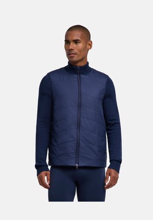 Mężczyzna w granatowej pikowanej kamizelce na suwak, na dopasowanym swetrze z długim rękawem, na tle jednolitego jasnego tła.